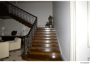 This photograph shows the interior of a luxurious residence, depicting a grand wooden staircase with ornate wrought-iron railings, polished hardwood steps, and elegant architectural details. The image includes period furniture and decorative objects 