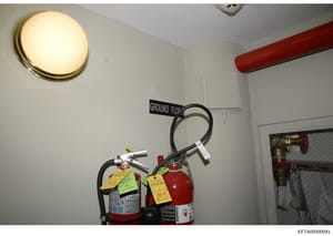 This photograph documents fire safety equipment located on the ground floor of one of Epstein's properties. The image shows two fire extinguishers mounted on a wall, labeled with inspection tags dated 2018, suggesting routine maintenance records. The