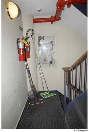This photograph shows an interior stairwell or landing area, likely from one of Epstein's properties, featuring fire suppression equipment (fire extinguishers and red piping), emergency lighting, cleaning supplies, and a wooden staircase railing. The