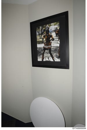 This is a photograph of a framed artwork hanging on a white interior wall. The framed image depicts a person in traditional African dress with beaded accessories and body paint, photographed outdoors with trees and white buildings visible in the back