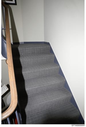 This is a photograph of an interior staircase, likely from one of Epstein's properties documented during the investigation. The image shows a flight of stairs covered with gray textured carpet, a wooden handrail, and white walls. The photo appears to