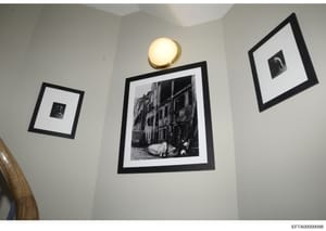 This photograph documents the interior decoration of a room, showing a white wall with three black-framed artworks and a brass wall sconce. The central and largest frame contains a black and white photograph of a European street scene with classical 
