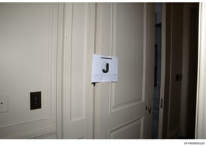 This is an evidence photograph from the Epstein investigation showing an interior room with cream-colored paneled walls, a dark electrical panel on the left, and a large white evidence marker labeled 'J' mounted on the wall. The photo appears to be p