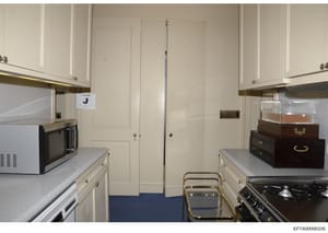 This is an evidence photograph of a kitchen interior, labeled with marker 'J' for identification purposes. The image shows a galley-style kitchen with cream-colored cabinetry, stainless steel appliances, and a blue floor. The photo appears to be part