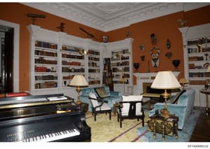 This is a high-quality interior photograph of an elegantly decorated library or study room featuring extensive built-in bookshelves, a grand piano, upholstered seating, decorative artifacts, and classical architectural details. The room displays sign