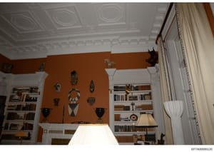 This photograph shows the interior of an upscale residential room featuring burnt orange walls, ornate white ceiling with decorative molding, built-in bookshelves with leather-bound volumes, and a curated collection of African masks and sculptures di