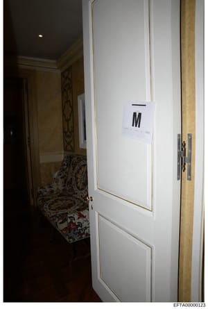 This is an evidence photograph from the Epstein investigation showing an interior room or hallway with a white door bearing an evidence marker labeled 'M'. The image captures hallway architecture with wood paneling, decorative elements, and ornate fu