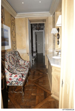 This photograph shows a luxuriously appointed hallway with adjoining bathroom in what appears to be one of Jeffrey Epstein's properties. The space features high-end finishes including travertine walls, decorative wall sconces, an ornate upholstered c