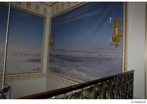 This photograph shows an ornate interior stairwell featuring large-scale cloud murals painted on walls, decorative gold-gilded wall sconces, ornamental crown molding, and an elaborate black iron railing. The scene depicts luxurious residential interi
