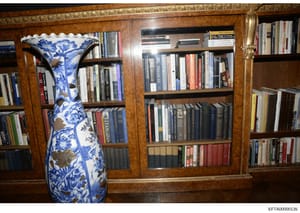 This photograph shows the interior of a residence, depicting a large wooden bookcase filled with books and a prominent blue and white porcelain decorative vase. The image appears to be evidence documentation from the Epstein investigation, likely tak
