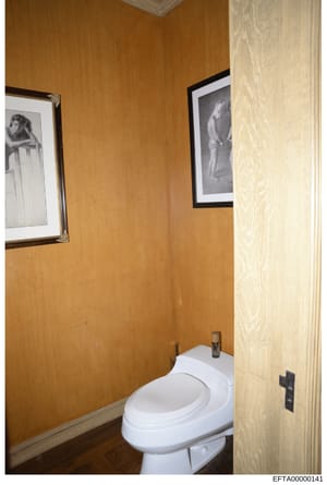 This is a photograph of a bathroom interior, showing wood-paneled walls in warm tones, a white toilet, and two framed black-and-white photographs hung on the walls. The image appears to be evidence documentation from a property search or seizure rela