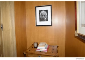 This photograph shows the interior of a room featuring wood-paneled walls, a wooden desk or table with books and a beverage cup, and a framed black-and-white portrait of an elderly man with distinctive white hair hanging on the wall. The image appear