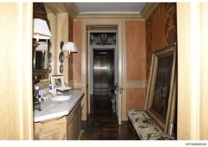 This is a photograph of an elegantly appointed bathroom and adjoining hallway from what appears to be a high-end residential property. The image shows custom marble countertops, brass fixtures, damask wallpaper, ornate mirrors, and decorative furnish