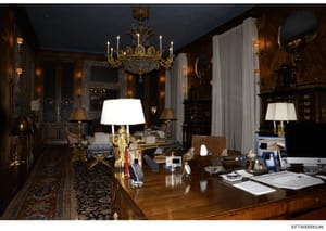 This is a photograph of an elegantly decorated interior room featuring dark wood paneling, a crystal chandelier, gold-trimmed mirrors, and luxurious furniture. The space appears to be a formal study or office with a large wooden desk displaying docum