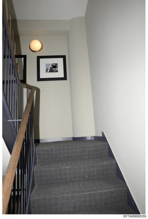This photograph shows an interior stairwell and hallway from an Epstein property, featuring a modern architectural design with white walls, black-framed artwork, wooden railings, and contemporary lighting fixtures. The image appears to be evidence do