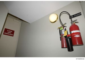 This is an evidence photograph from an Epstein property showing a third-floor hallway with a prominent '24 HOUR VIDEO SURVEILLANCE' sign on a door, a ceiling-mounted light fixture, and fire safety equipment including multiple fire extinguishers mount
