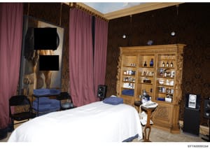 This is a photograph of an interior room, likely a massage parlor or spa treatment room, showing furnishings and equipment including a treatment table, wooden cabinet with various bottles and products, curtained areas, and audio equipment. The room f