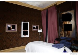 This photograph depicts an interior bedroom space featuring ornate décor, including damask-patterned wallpaper in brown and gold tones, burgundy curtains, framed artwork with redacted content, a white bed with blue cushions, and various furnishings. 