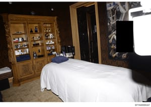 This is an evidence photograph depicting an interior bedroom space containing luxurious furnishings, including an ornate wooden cabinet system with shelving displaying various personal items and toiletries, a bed with white linens, and decorative ele