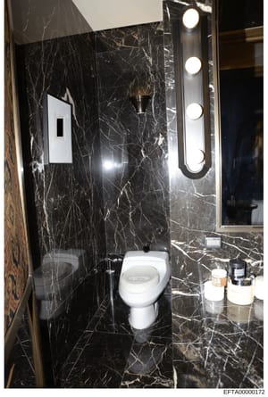 This is a photograph of a luxury bathroom interior, featuring high-end finishes including black marble walls with gold veining, a white ceramic toilet, illuminated vanity mirror with multiple light fixtures, and bathroom accessories on the counter. T