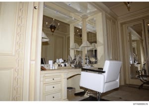 This is a photograph of an elegantly appointed bathroom or dressing room featuring a custom-built vanity with extensive mirror panels, ornate molding, and high-end finishes. The space displays cream-colored cabinetry, decorative elements including sc