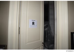 This is a photograph of an interior room showing a cream-colored wall with vertical molding and a doorway. A white evidence marker labeled 'R' is positioned on the wall. A black plastic evidence bag is visible in the doorway behind the marker. The ph