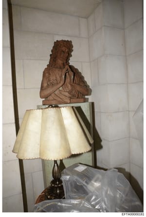 This photograph shows a terracotta or clay bust sculpture mounted on a pedestal with a cream-colored fabric lampshade, positioned in a corner room with white tile walls. The image appears to be an inventory or evidence photograph documenting property