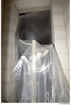 This is an evidence photograph showing a plastic-wrapped object stored in what appears to be a narrow ceiling or wall cavity. The item is wrapped in clear plastic sheeting and appears to be a cylindrical or tubular object. The photograph was taken as
