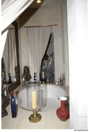 This is a photograph of an upscale bathroom interior, showing a marble countertop with decorative items including a brass candle holder, bronze bust sculpture, colored glass bottles, and a red ceramic vessel. The image documents the interior design a