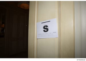 This is an evidence photograph showing an interior room with cream-colored walls and a dark wooden door on the left. A white evidence marker labeled with a large 'S' is affixed to the wall, along with what appears to be a document reference form or e