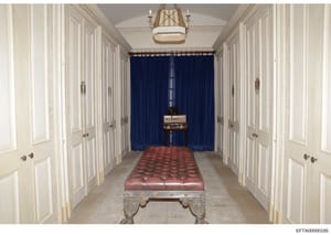 This is a photograph of a long, formal hallway in what appears to be a luxury residence, featuring white paneled closet doors on both sides, a red upholstered bench in the center, deep blue curtains at the far end, and decorative architectural elemen