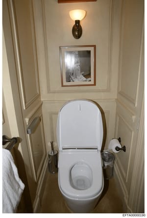 This is an evidence photograph showing a bathroom interior with a white toilet, wall sconces, framed artwork, and decorative fixtures. The image appears to be documentation of property interior spaces, likely taken during the investigation of one of 