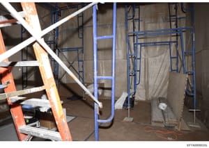 This photograph shows an interior space under active construction or renovation, featuring blue metal scaffolding, protective plastic sheeting, an orange ladder, and construction equipment. The image appears to be documentation of a property renovati