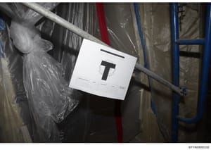 This is an evidence photograph showing a packaged item in storage marked with an evidence tag labeled 'T'. The item appears to be wrapped in plastic and stored alongside other materials on what looks like shelving or storage racks. The evidence marke