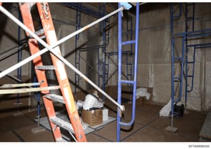 This is an evidence photograph showing the interior of what appears to be a property under construction or renovation. The image depicts blue metal scaffolding, an orange-red ladder, white piping, plastic sheeting, cardboard boxes, and various constr