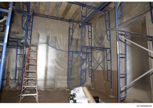 This photograph shows an interior space under construction or renovation, featuring blue metal scaffolding, protective plastic sheeting, and a red ladder. The image appears to be documentation of property improvements or renovation work, possibly at 