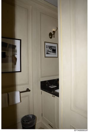 This is a photograph of a bathroom interior showing white paneled walls, black marble countertop, framed artwork, brass fixtures, and a towel rack. The image appears to be documentation of a property interior, likely from one of Jeffrey Epstein's res