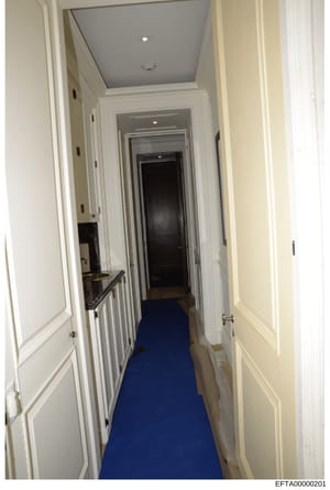 This is a photograph of an interior hallway in one of Jeffrey Epstein's properties, showing a narrow passage with white-painted walls, recessed ceiling lighting, and a distinctive blue carpet runner. The hallway connects multiple rooms via white-trim