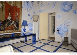 This photograph shows an elegantly decorated interior room featuring blue and white floral wallpaper, Asian-inspired artwork, and contemporary furnishings. The room contains a red lacquered panel with figurative artwork on the left, a blue table lamp