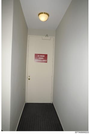 This is an evidence photograph showing a narrow interior hallway with white walls, a black ribbed floor mat, and a brass-handled white door. A prominent red sign reading '24 HOUR VIDEO SURVEILLANCE' is affixed to the door. The image documents securit