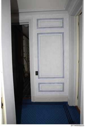 This is a photograph of a residential hallway showing a white paneled door with blue-gray trim and a blue patterned carpet. The image appears to be from an interior property inspection or evidence documentation related to the investigation, showing a