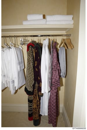 This photograph shows the interior of a bathroom or bedroom closet containing folded towels on an upper shelf, wooden hangers with various clothing items including white garments, patterned scarves or wraps in black and gold, burgundy, and pink tones