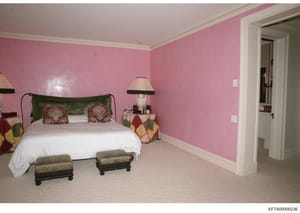 This is a photograph of a residential bedroom featuring distinctive pink walls, white ceiling, and ornate furnishings including a green wrought iron bed frame, decorative pillows, a bedside lamp, and two upholstered ottomans. The image appears to be 