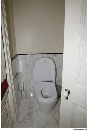 This is a photograph of a modern bathroom interior showing a white toilet with closed seat lid, marble flooring with gray veining, white painted walls, and stainless steel fixtures. The image appears to be documentation of property interior condition