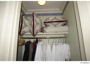 This is a photograph of an organized closet interior, likely from one of Jeffrey Epstein's residences. The image shows shelving with decorative pillows and linens with red velvet trim and blue floral patterns on the upper shelf, and a clothing rod be