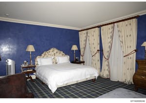 This is a photograph of a luxuriously appointed bedroom featuring deep blue walls, a decorative upholstered headboard, white bedding, cream-colored patterned curtains with burgundy tie-backs, wooden furniture, and a plaid carpet. The photograph appea