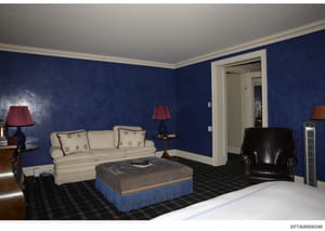 This is a photograph of an elegantly furnished bedroom featuring deep blue walls, cream-colored seating furniture, and formal décor. The room contains a cream sofa with decorative pillows, a black leather armchair, a striped ottoman with blue skirt, 