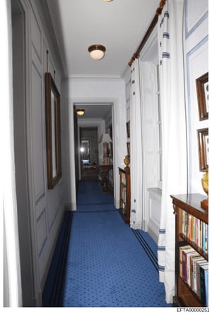 This is a photograph of a long, narrow hallway in what appears to be an upscale residential property, featuring white-paneled walls, a blue patterned carpet with decorative borders, wooden-framed artwork on the walls, built-in bookshelves, and classi