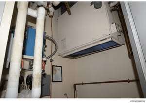 This photograph shows the interior of a utility or mechanical room, likely from one of Epstein's properties. The image captures various building systems including PVC piping, a blue water heater unit, copper piping, and ceiling-mounted equipment. The