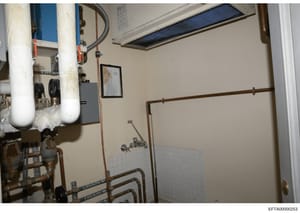 This photograph shows the interior of a utility/mechanical room, likely in a residential property, featuring water heaters, plumbing fixtures, HVAC equipment, and various pipes and controls. The image appears to be evidence documentation from a prope